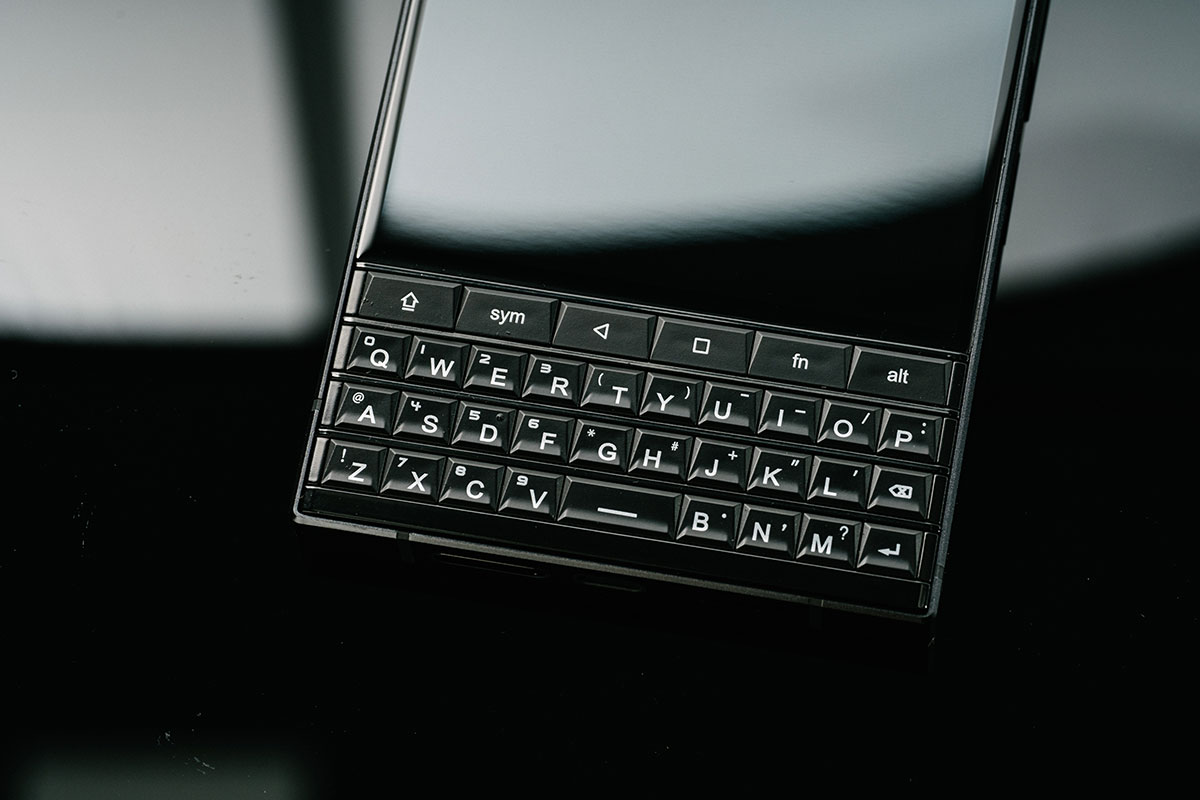 Смартфон із фізичною QWERTY-клавіатурою чорного кольору, розташований на темній глянцевій поверхні; видно компактні рельєфні клавіші та екран над ними.